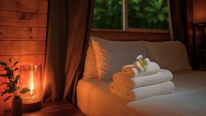 A neatly made bed with white linens and a folded towel, a vase with a flower, and a lit candle on a wooden nightstand.