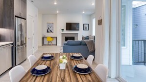 A modern kitchen with a built-in refrigerator, a dining area with a wooden table set for four, and a living room with a sofa and a flat-screen TV.