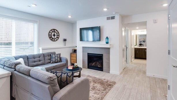 A modern living room with a grey sofa, a fireplace, a TV mounted above it, and a dining area visible through an open door.