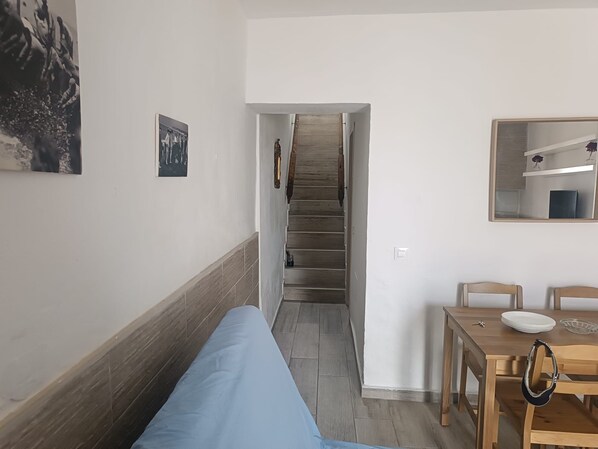 Un couloir avec un escalier, un coin repas avec une table et des chaises, et un lit recouvert d’une couverture bleue.