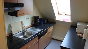 Une cuisine dotée d’un plan de travail noir, d’un évier en acier inoxydable, de meubles en bois, d’une machine à café et d’une fenêtre laissant entrer la lumière naturelle.