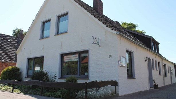 Un bâtiment blanc à deux étages, doté d’un toit à pignon, portant le numéro 34, avec de grandes fenêtres et un petit jardin clos par une clôture en bois.