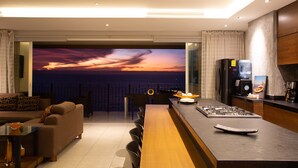 A modern living room with a large TV displaying a sunset view, a kitchen area with a gas stove, and a dining area with a bar. - ★ 15th Floor Grand Venetian ★ (Puerto Vallarta)