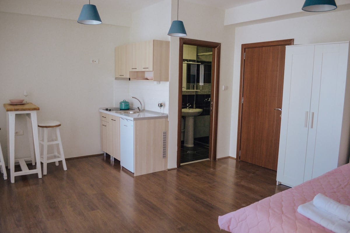 Une pièce compacte comprenant un coin cuisine, une armoire en bois et un lit recouvert d’une courtepointe rose.