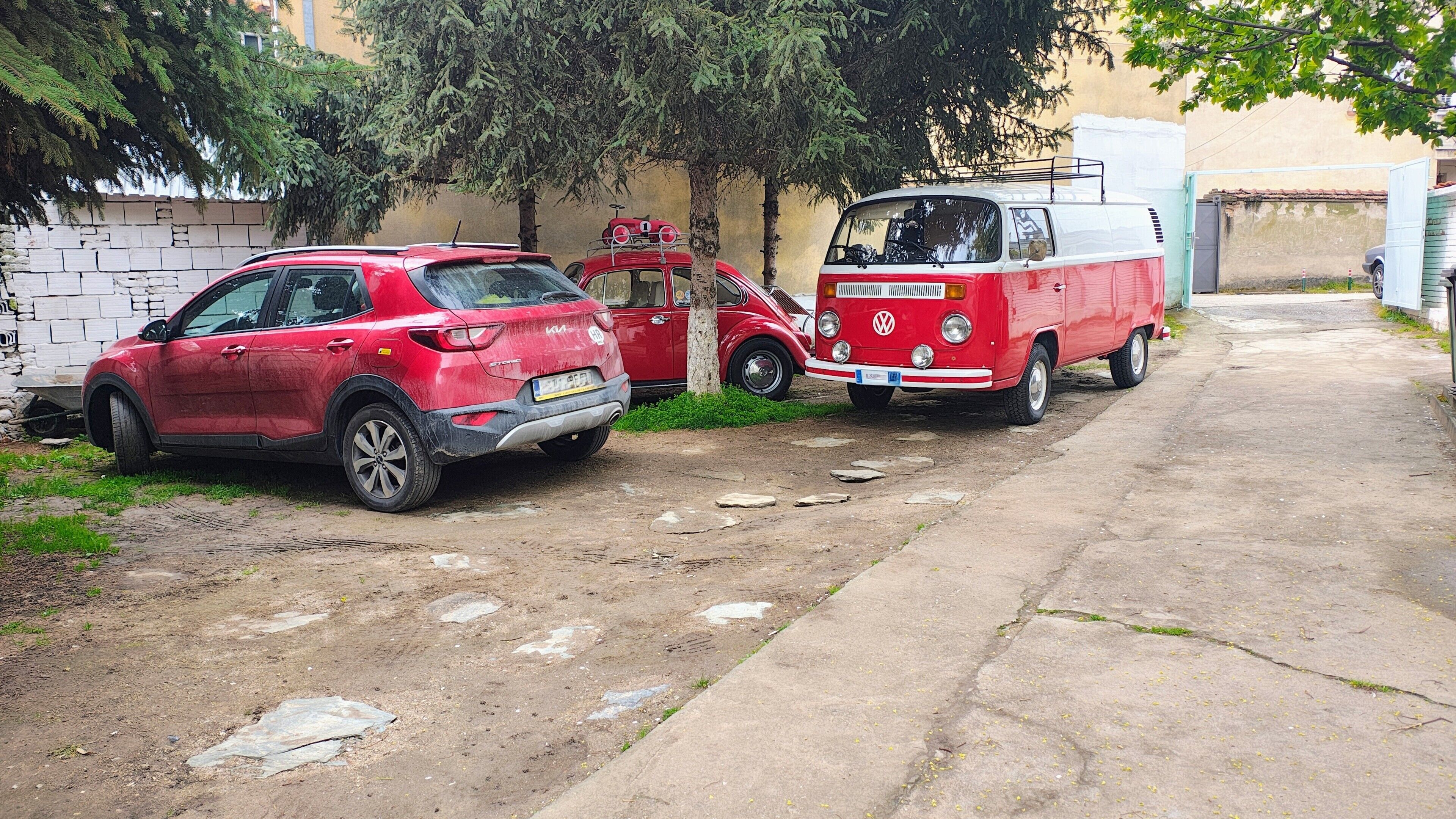 A red SUV and a vintage VW van parked on a dirt path beside a building with a stone wall.