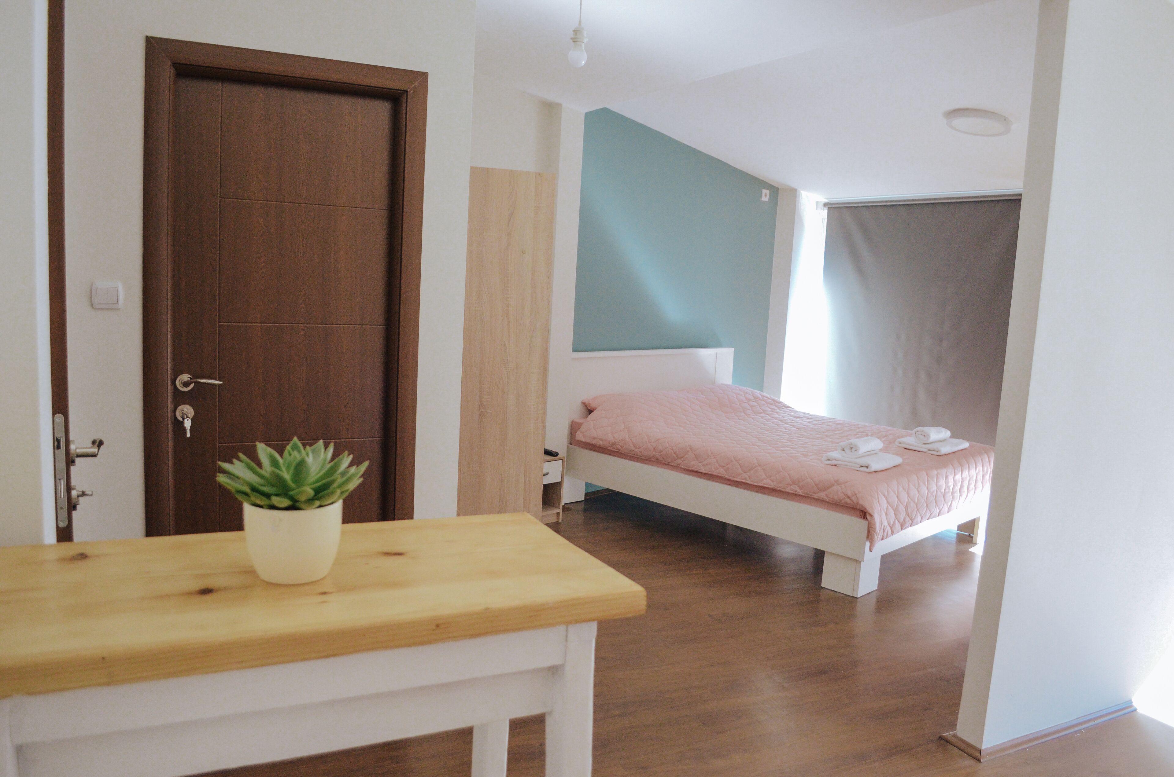 Une chambre avec un plancher en bois, un lit recouvert d’une couette rose, une table de chevet blanche ornée d’une plante, et une porte dotée d’une serrure à clé.