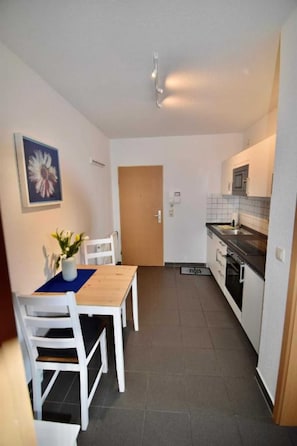 Une cuisine compacte avec un coin repas, comprenant une table en bois, des chaises blanches, une petite plante en pot et une porte donnant sur une autre pièce.