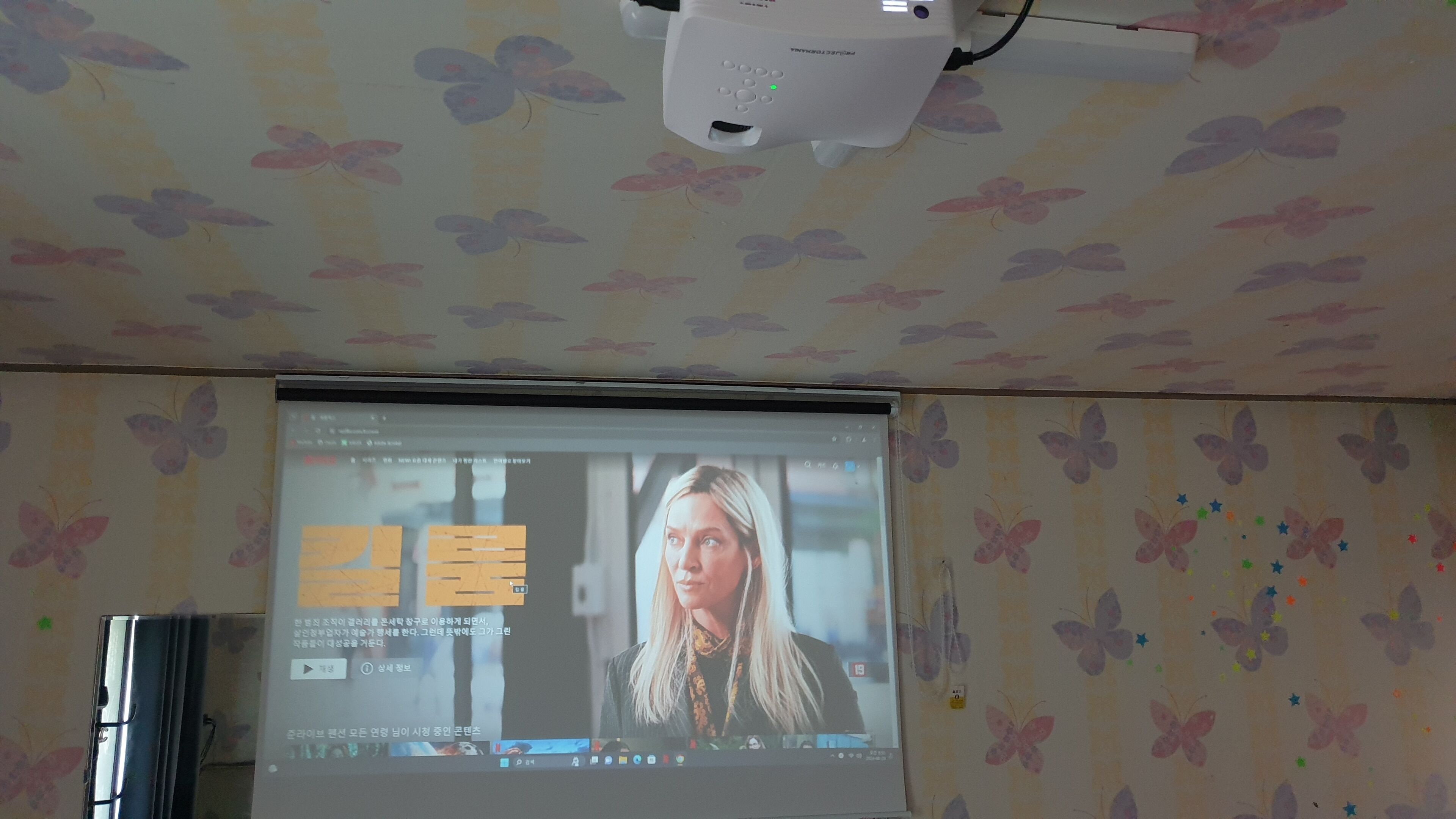Pantalla de proyección mostrando un video con una persona, un proyector y una pared con diseño.