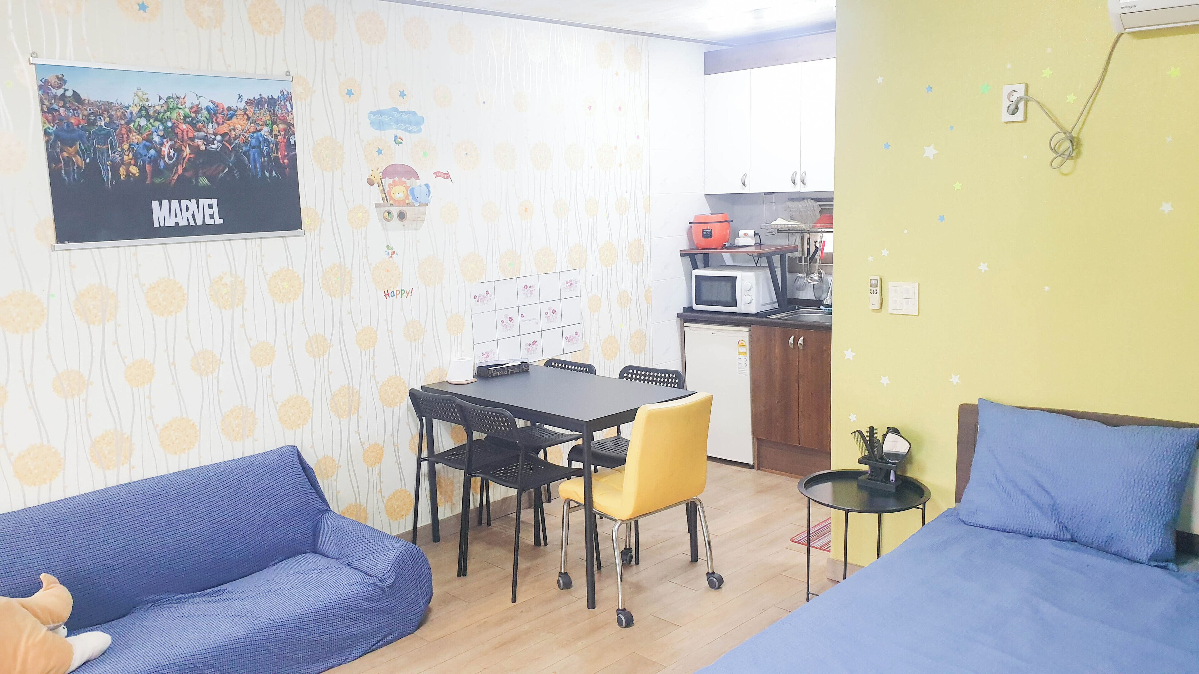 A small, cozy room with a blue sofa, a yellow chair, a small table, and a bed. There is a kitchen area with a microwave and a sink.
