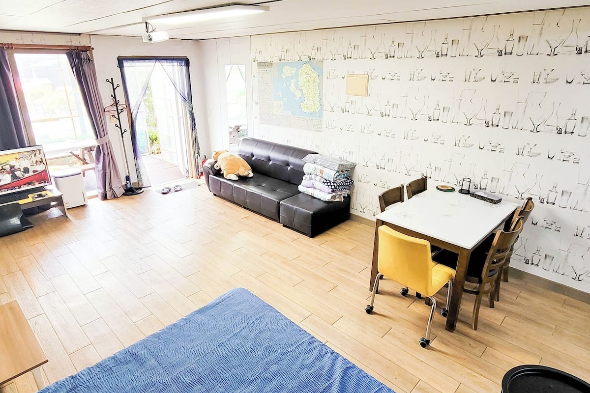 A living room with a sofa, a table, and a chair. There is a window with curtains, a wall with a map, and a door leading outside.