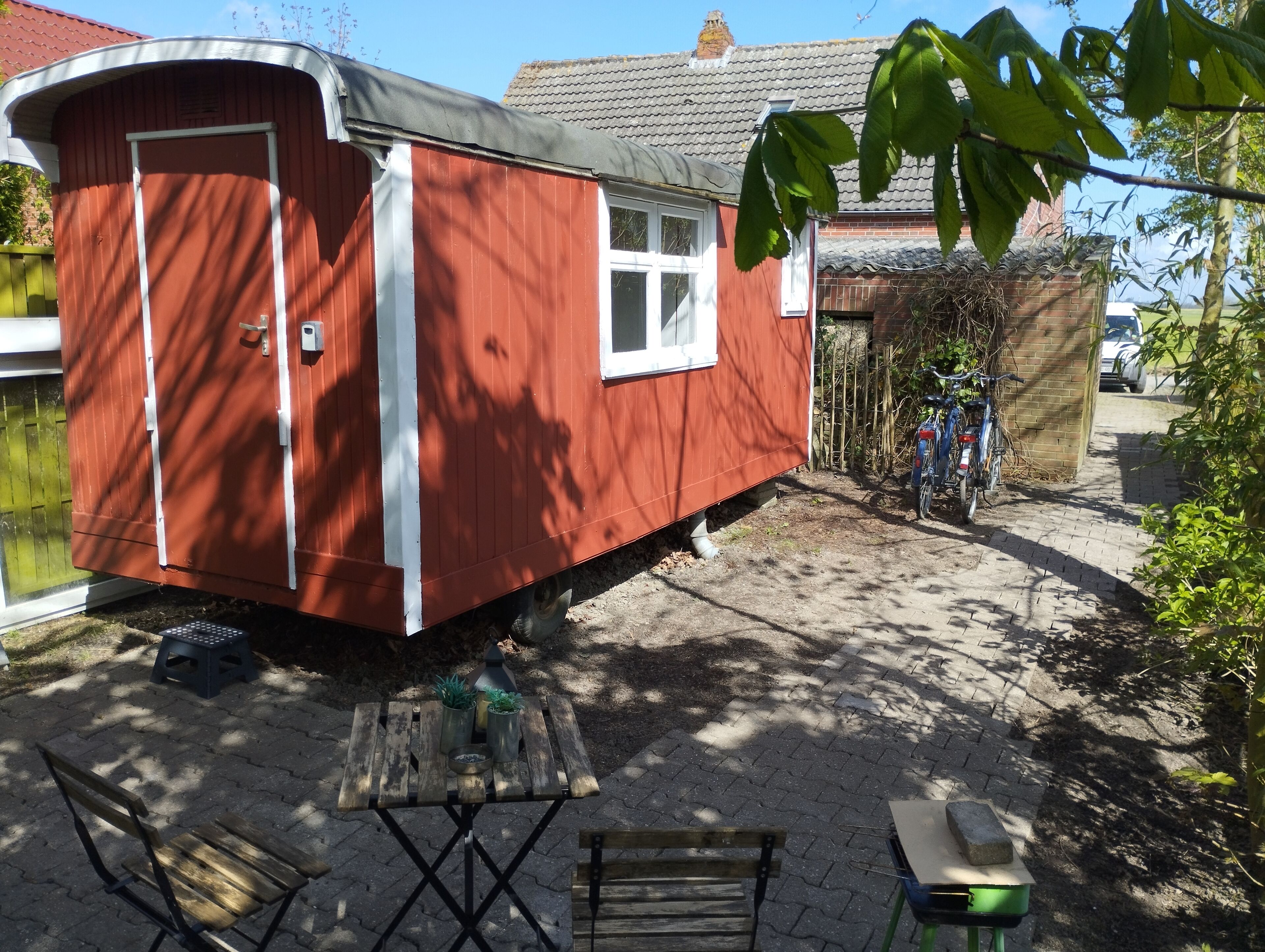 A red mobile home with a white window and door, a small patio area with a table and chairs, and a bicycle parked nearby.