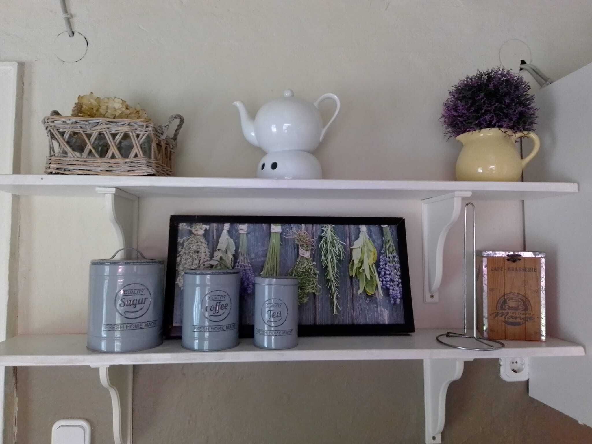 En hylde med en hvid tepotte, en gul kande, et indrammet billede af krydderurter, og tre sølvbeholdere med teksten "Quality Coffee" og "Quality Tea".