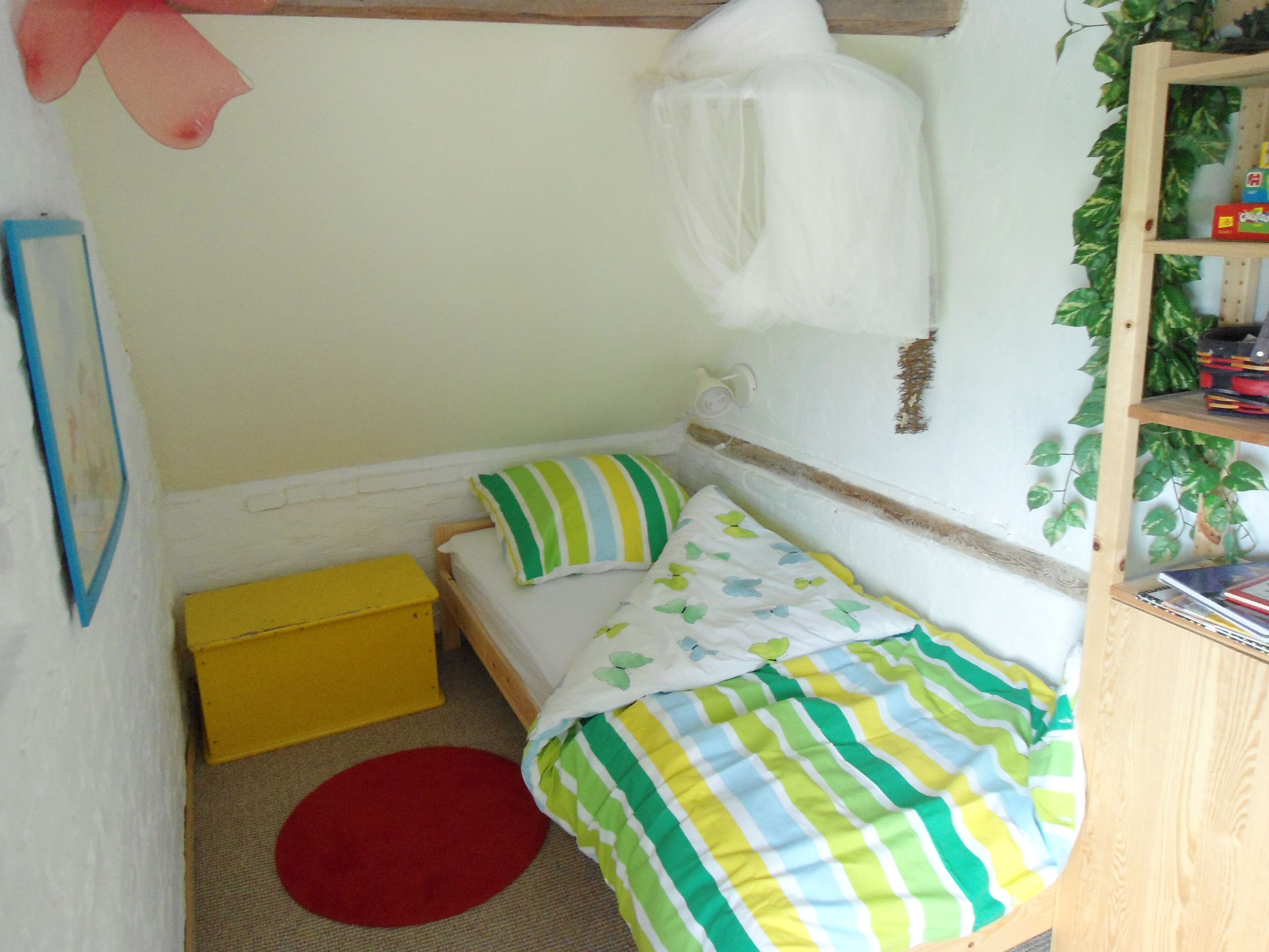 Een knus slaapkamer met een enkel bed, een gele opbergkist, een rode vloerkleed en een houten plank met boeken.