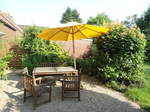 Ein Holztisch mit Stühlen steht unter einem gelben Sonnenschirm in einem Garten mit Kiesböden, umgeben von Grünpflanzen und einer Backsteinmauer.