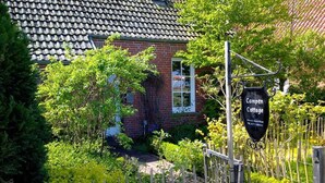 Un bâtiment en briques surmonté d’un toit en tuiles, une enseigne indiquant « Camden Cottage », et une clôture en bois agrémentée de verdure.