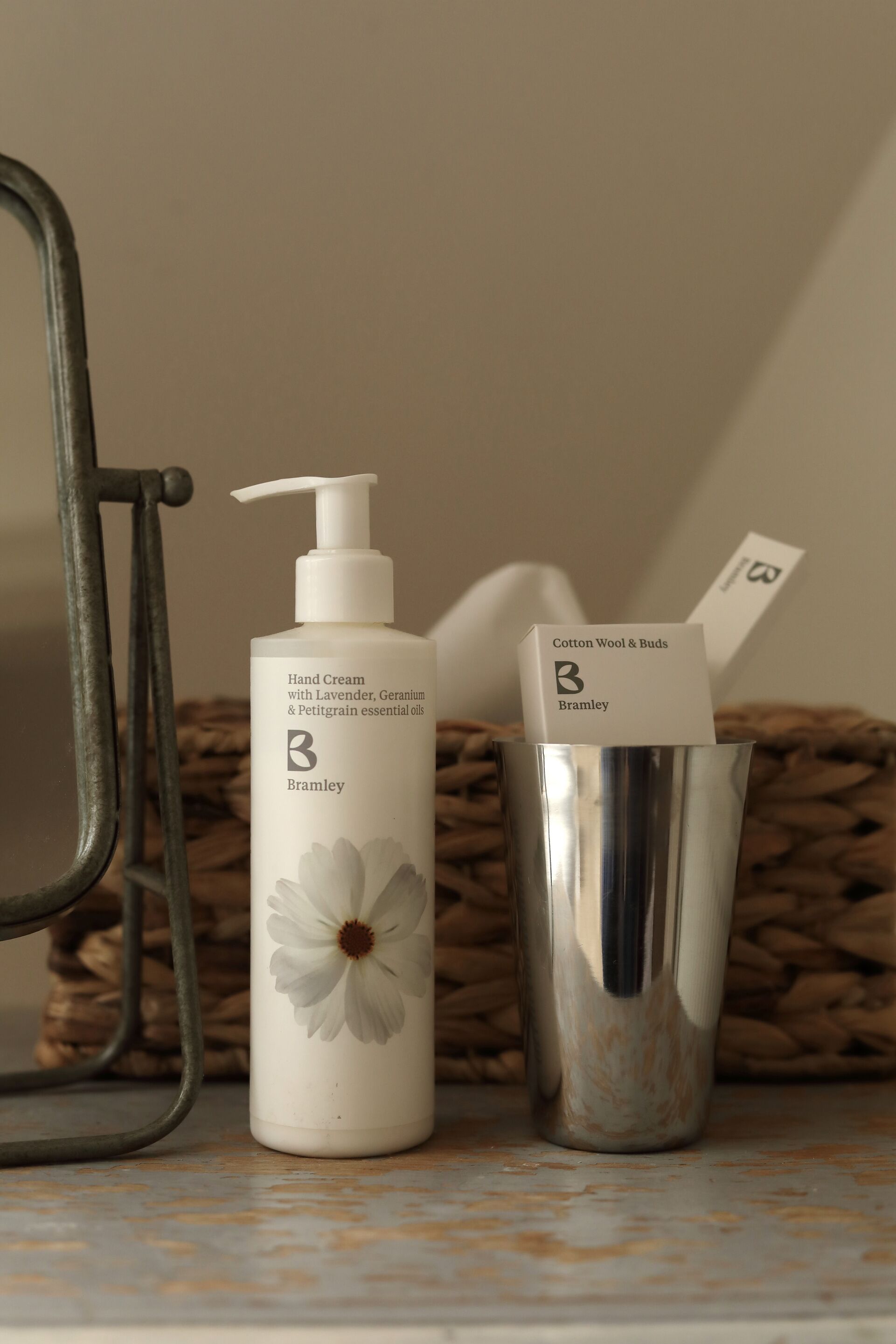 Een handcrème fles met een bloemenetiket, een metalen beker met een kaartje, en een rieten mand op de achtergrond.