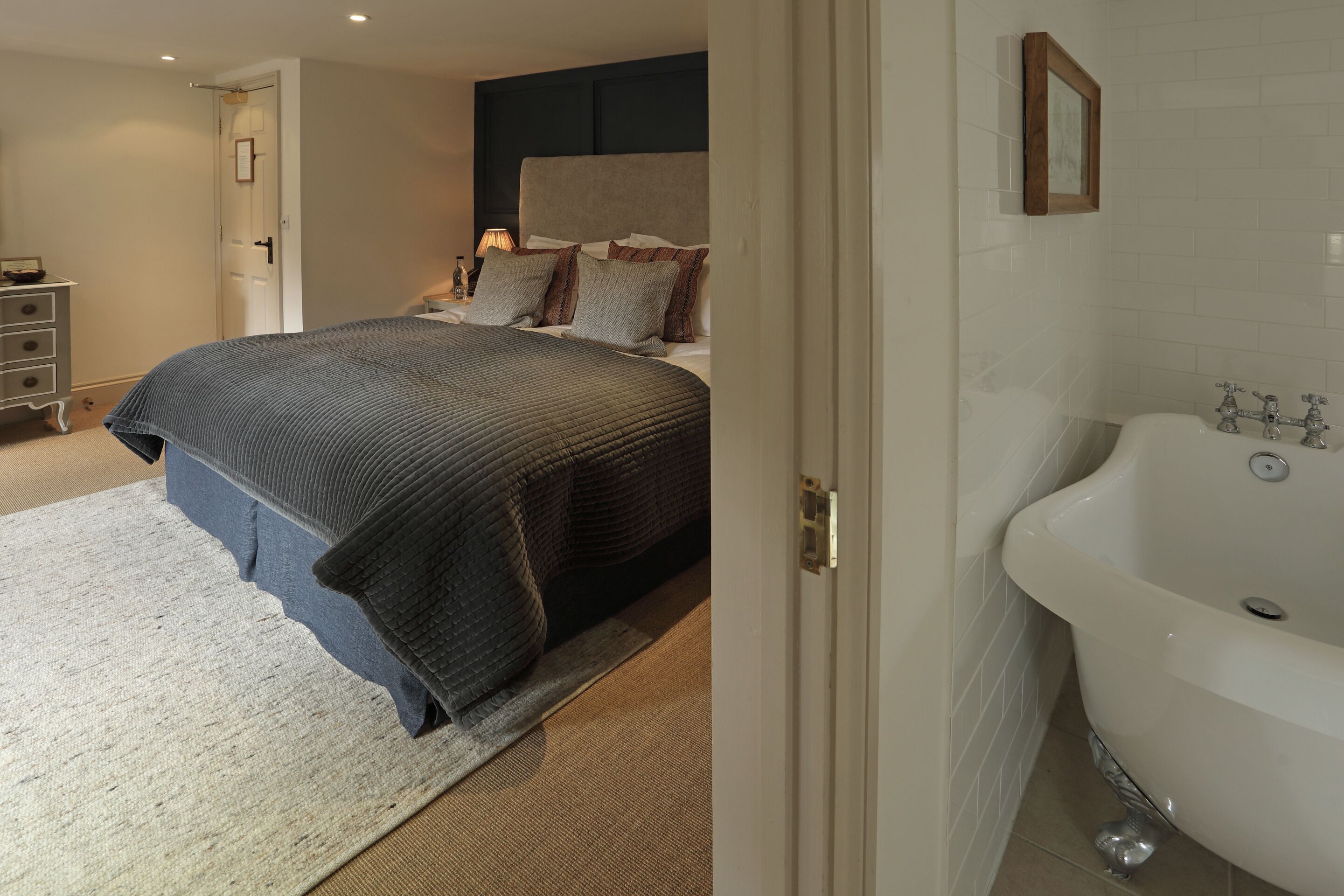 A bedroom with a large bed, a bedside table, a lamp, and a view of a bathroom with a bathtub.