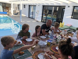 A family dining outdoors by a pool with a modern house in the background. - Brand new and modern villa in Lajares (Fuerteventura) with heated pool (Lajares - Fuerteventura)
