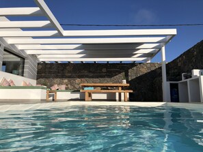 A modern outdoor pool area with a stone wall, a wooden table, and colorful cushions. - Brand new and modern villa in Lajares (Fuerteventura) with heated pool (Lajares - Fuerteventura)