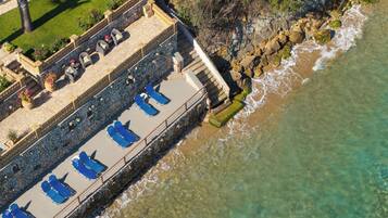 An aerial view of a coastal hotel with a stone deck, blue lounge chairs, and a stone wall overlooking the sea.