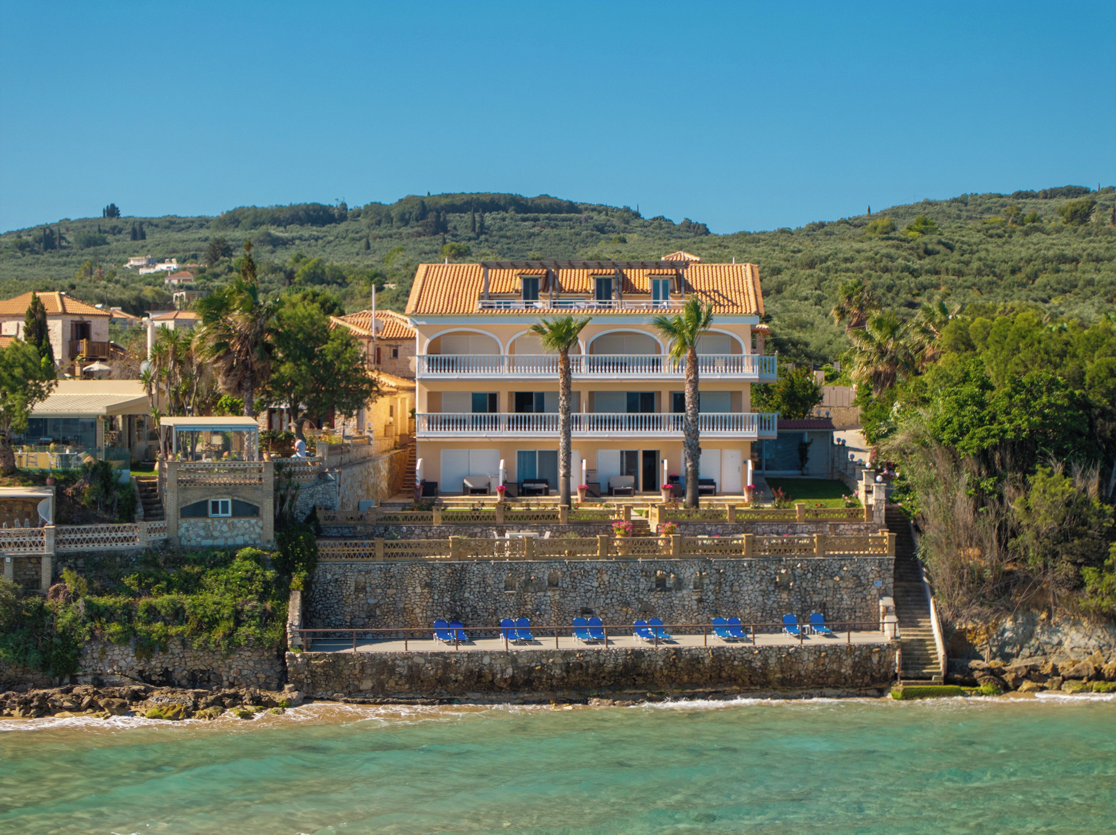 Un hôtel situé sur la côte, doté d’une façade en pierre, de nombreux balcons et d’un espace piscine avec des chaises bleues.
