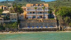 Un hôtel situé sur la côte, doté d’une façade en pierre, de nombreux balcons et d’un espace piscine avec des chaises bleues.