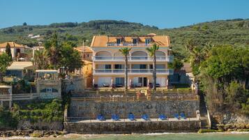 Un hôtel situé sur la côte, doté d’une façade en pierre, de nombreux balcons et d’un espace piscine avec des chaises bleues.
