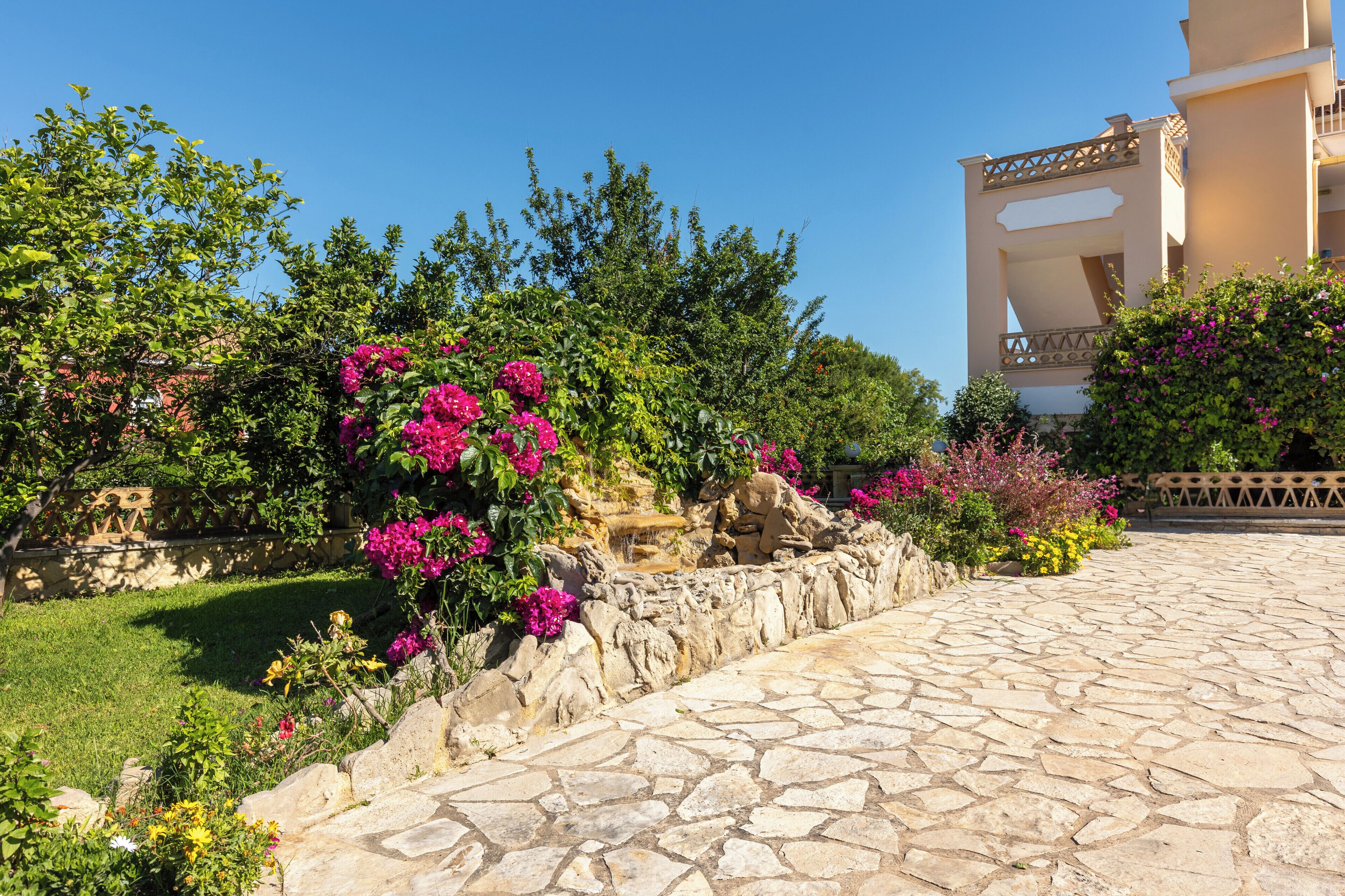 A stone pathway leading to a building with a balcony, surrounded by lush greenery and flowering plants.
