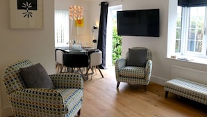 Een woonkamer met gestreepe fauteuils, een eettafel met stoelen, een aan de muur bevestigde televisie en een raam met uitzicht op groen.