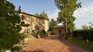 Un cortile con lastricato, un edificio in pietra con persiane rosse e vegetazione circostante.