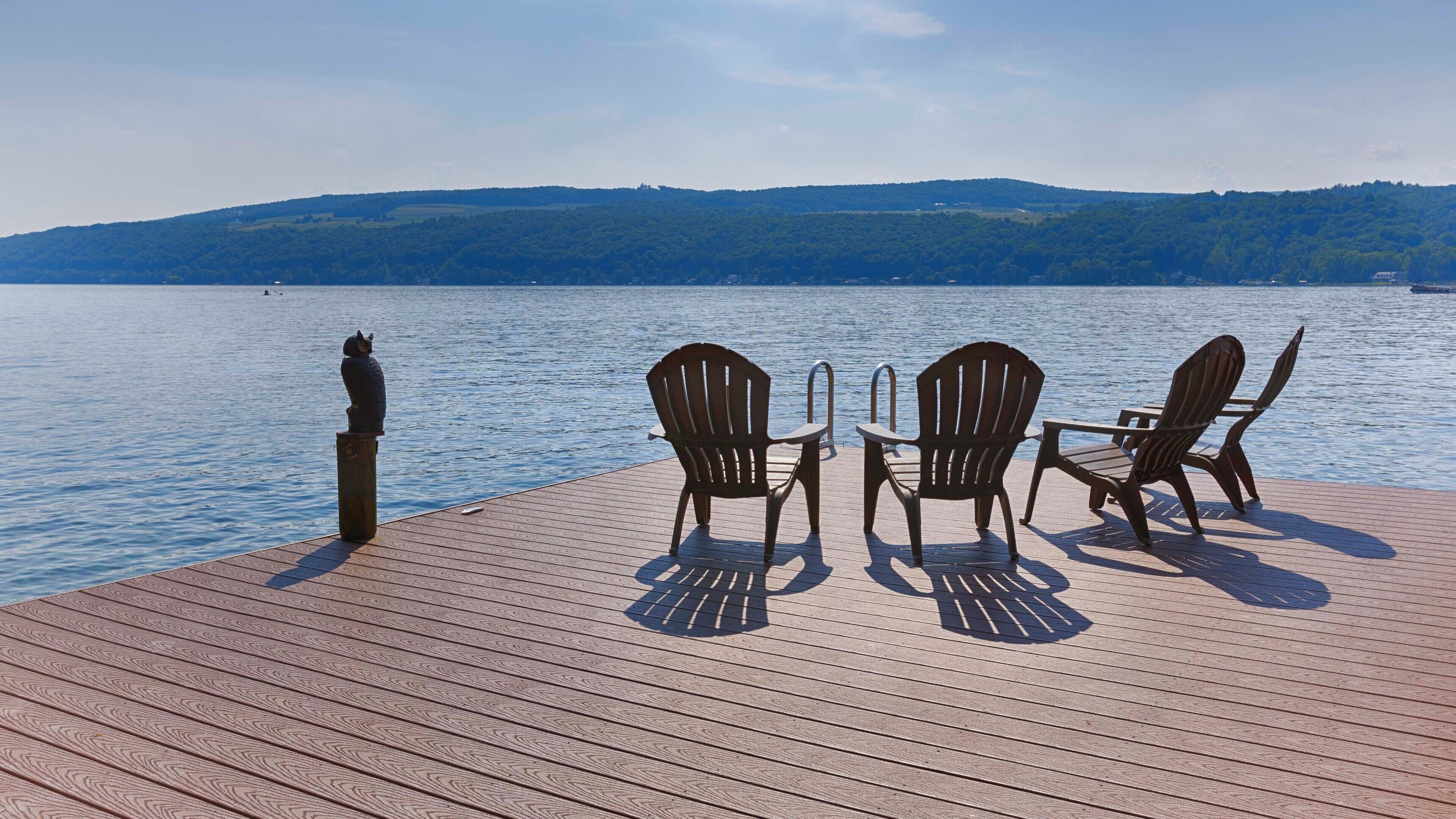 Eine Holzterrasse mit drei Adirondack-Stühlen und einem Tisch, mit Blick auf einen ruhigen See und entfernte Hügel.