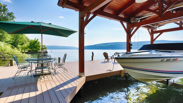 A wooden deck with a table and chairs, a covered area with an umbrella, and a boat docked at the pier. - Ironwood: Amazing Keuka Lake Views! (Hammondsport)
