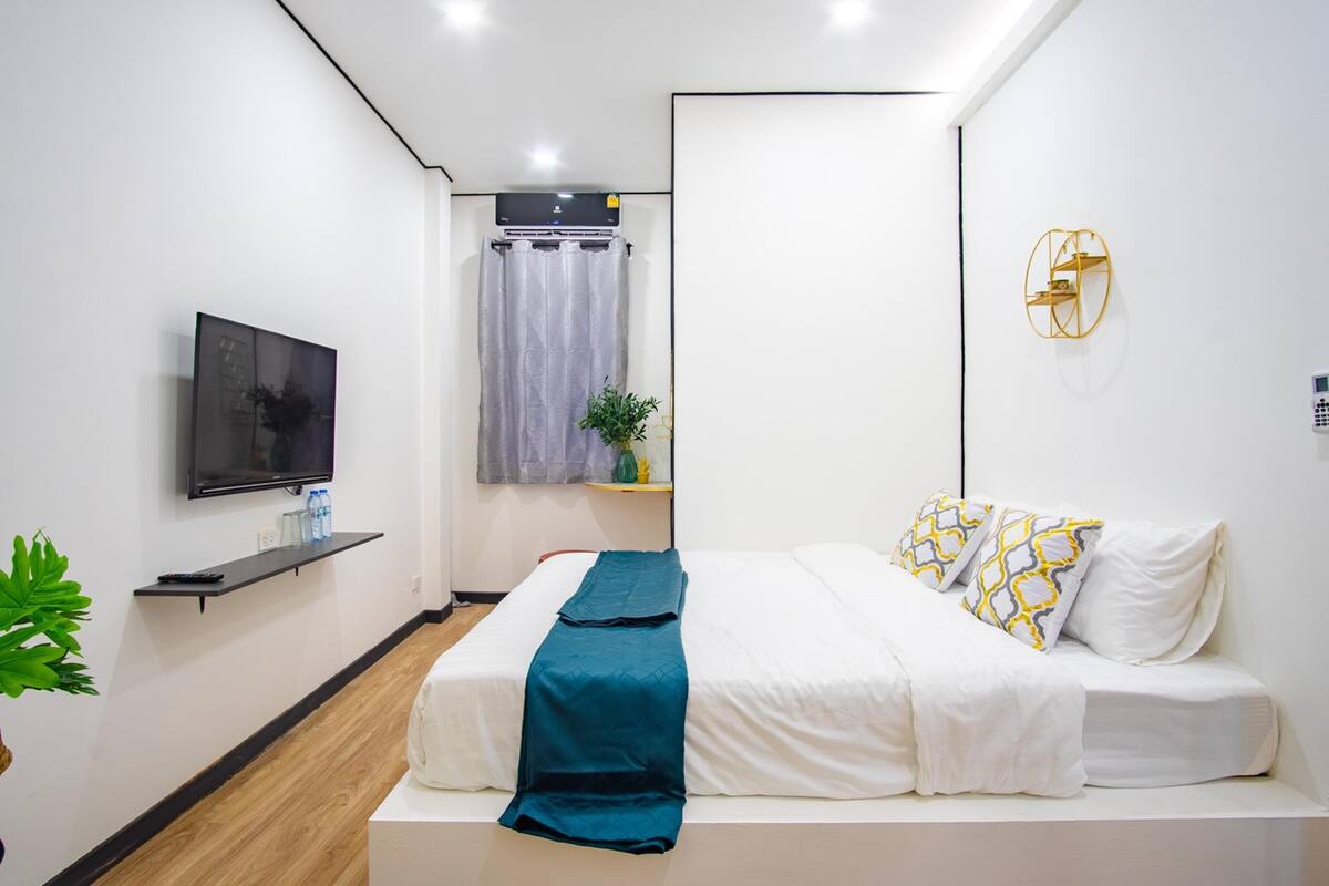 Habitació d'hotel moderna amb llit, televisió, finestra amb cortines, una planta i una estant penjada a la paret.