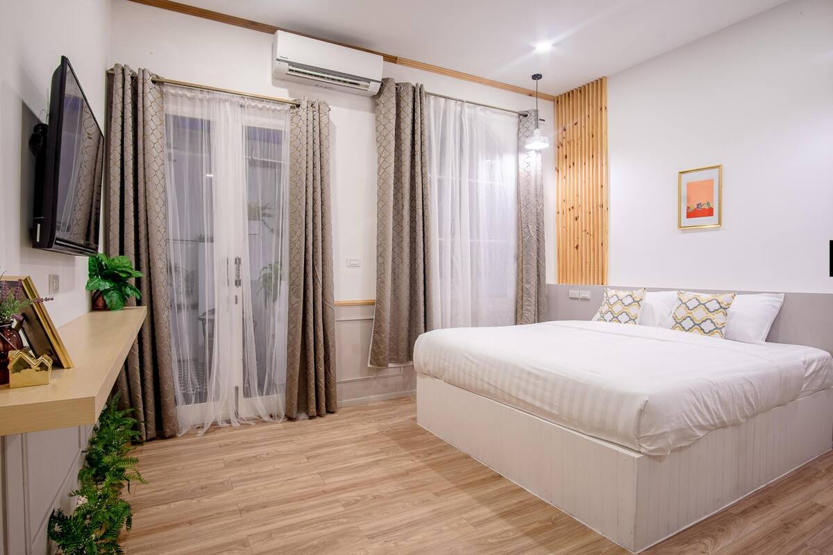 Un dormitori amb un llit, cortines, un televisor, una planta i un quadre penjat en una estantàlia.