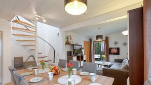 A modern dining area with a wooden table set for six, surrounded by grey upholstered chairs. A staircase with wooden steps and a glass railing leads to an upper level. - Cottage near Diane (lake, forest, wellness) (Kirchheim)