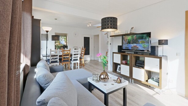 A modern living room with a sofa, coffee table, TV, and a dining area in the background. - Cottage near Diane (lake, forest, wellness) (Kirchheim)
