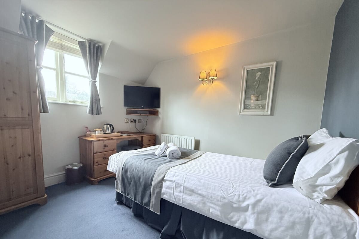 A bedroom with a bed, a desk, a chair, a television, a radiator, and a window with curtains.