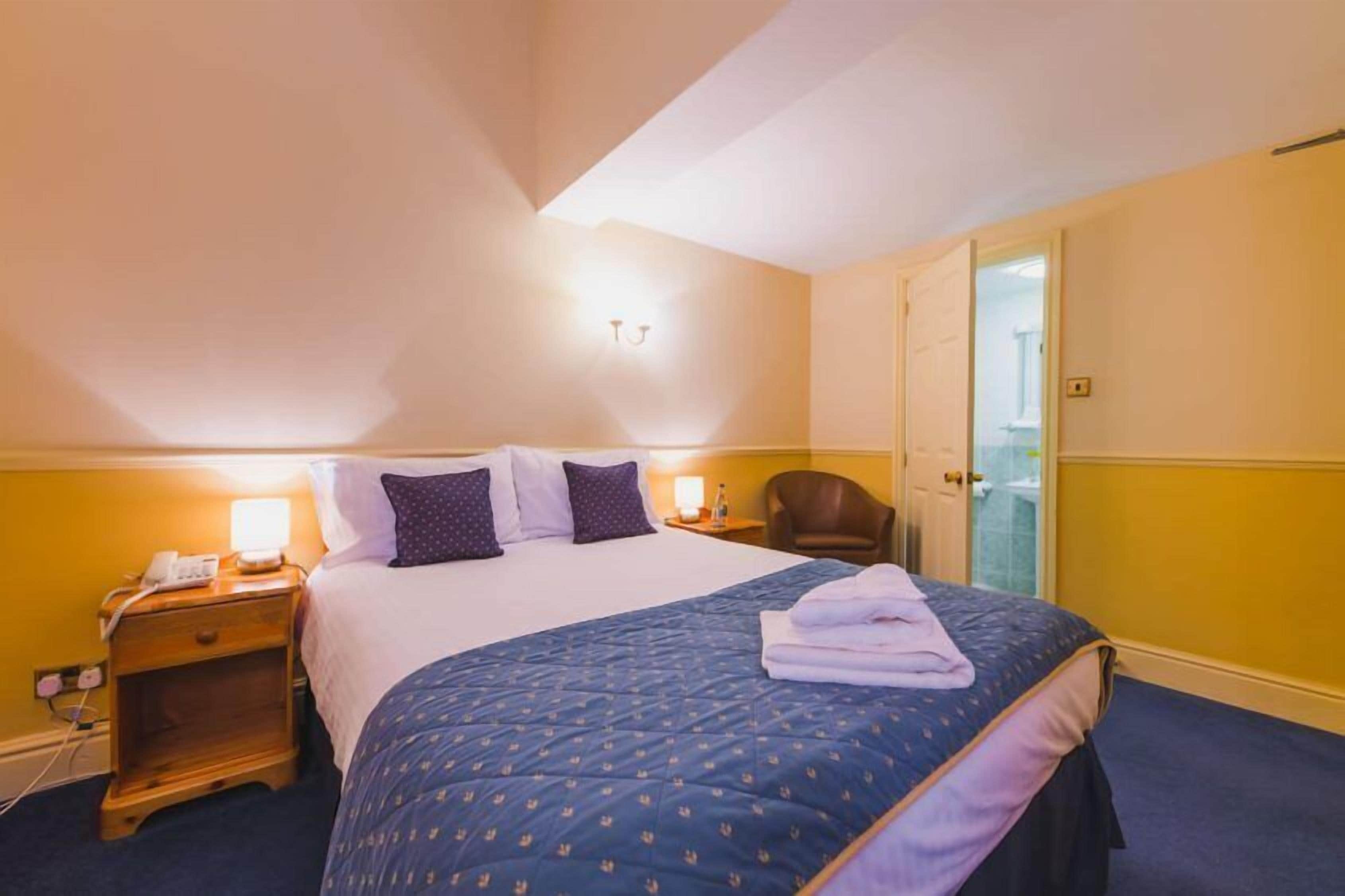Una habitación de hotel con una cama, mesitas de noche, una silla y una puerta que da a un baño.