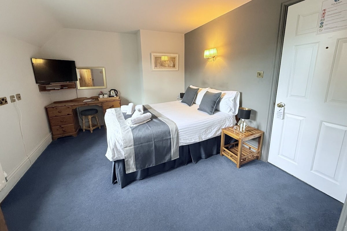 A hotel room with a bed, a desk, a chair, a television, and a door with a sign.