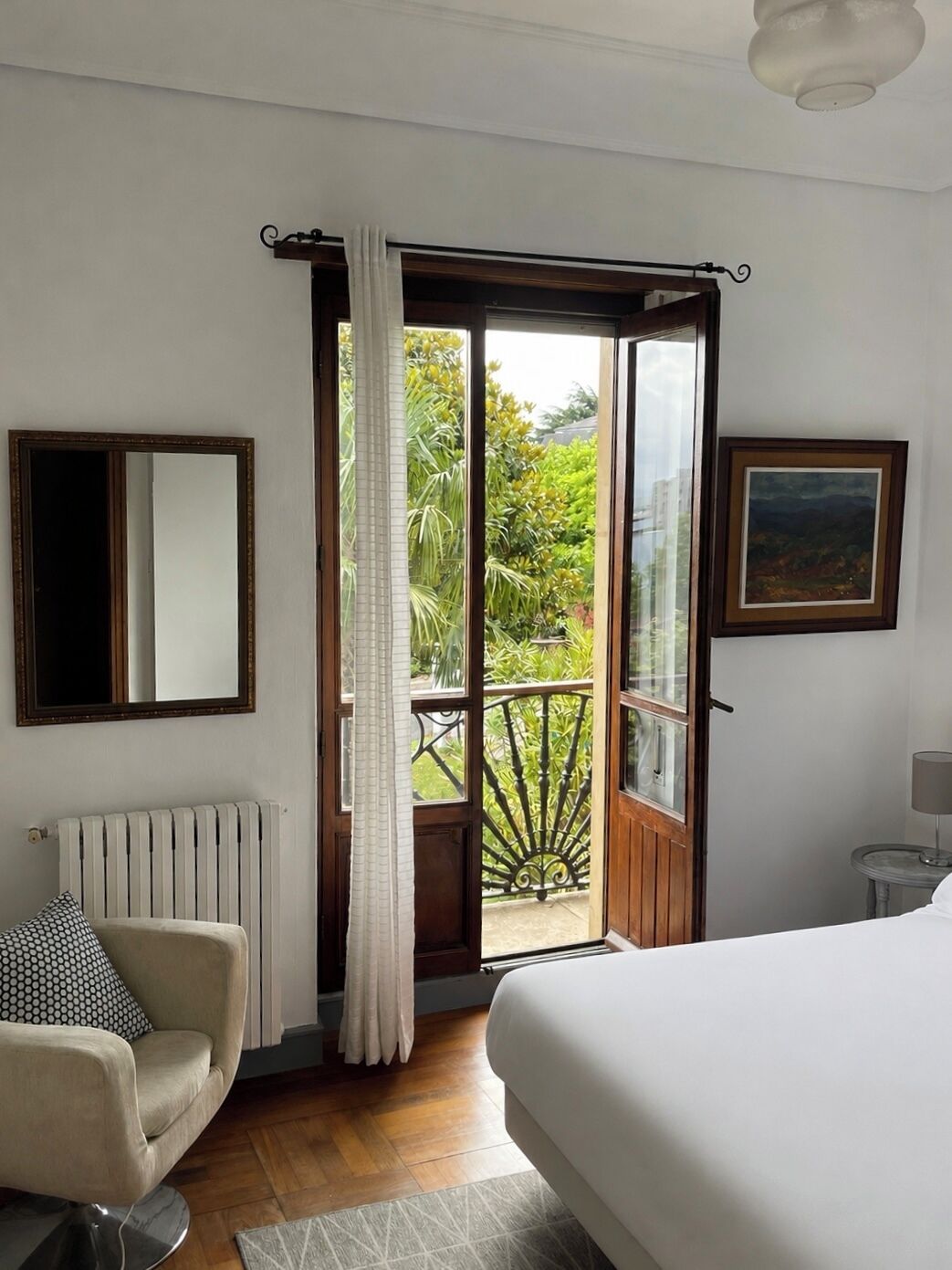 A bedroom with a bed, a chair, a radiator, a window with curtains, and a painting on the wall.