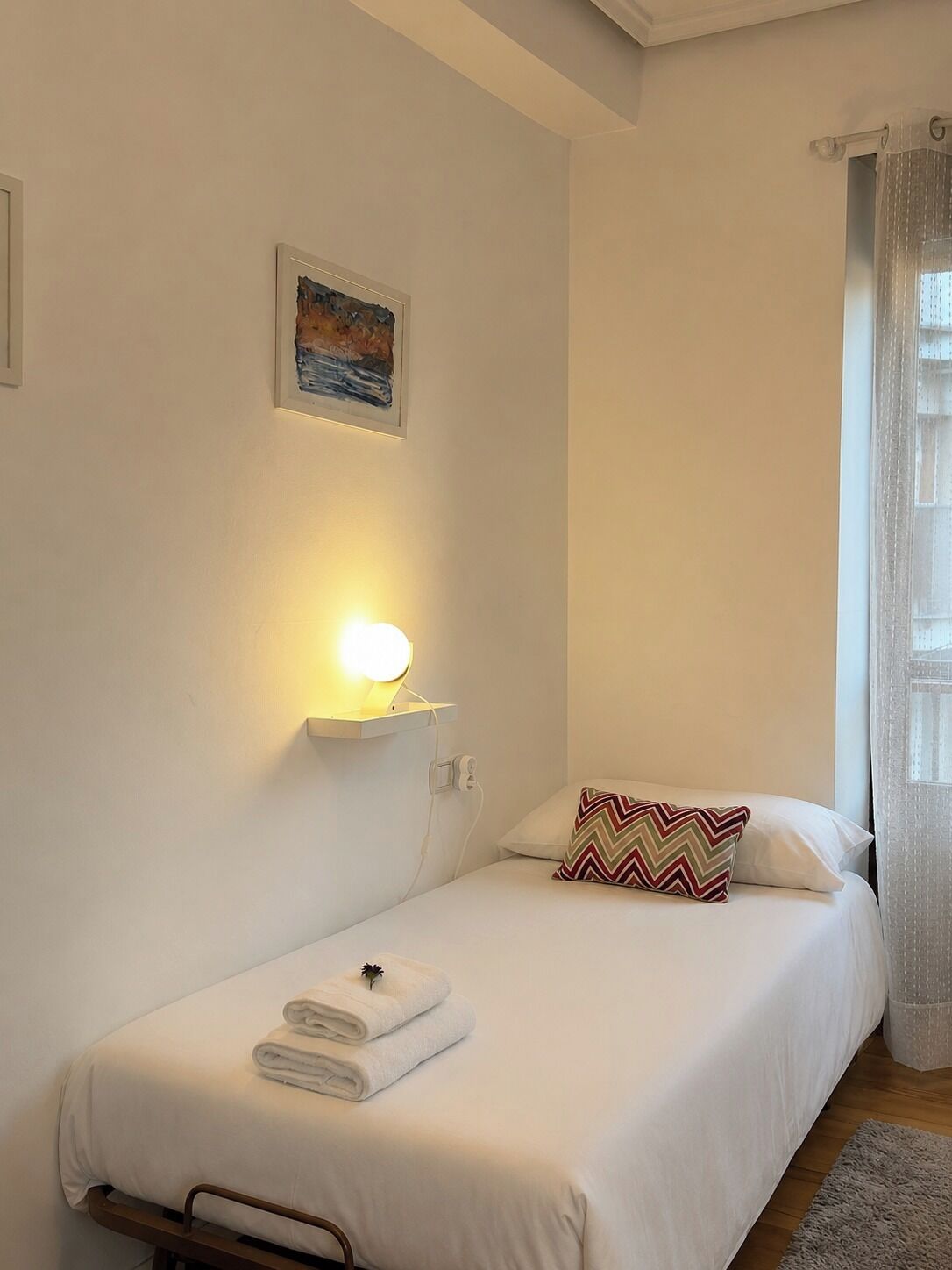 Una cama individual con almohada, una lámpara de pared, un cuadro y una ventana con cortinas.