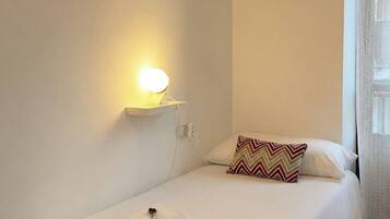 Una cama individual con almohada, una lámpara de pared, un cuadro y una ventana con cortinas.