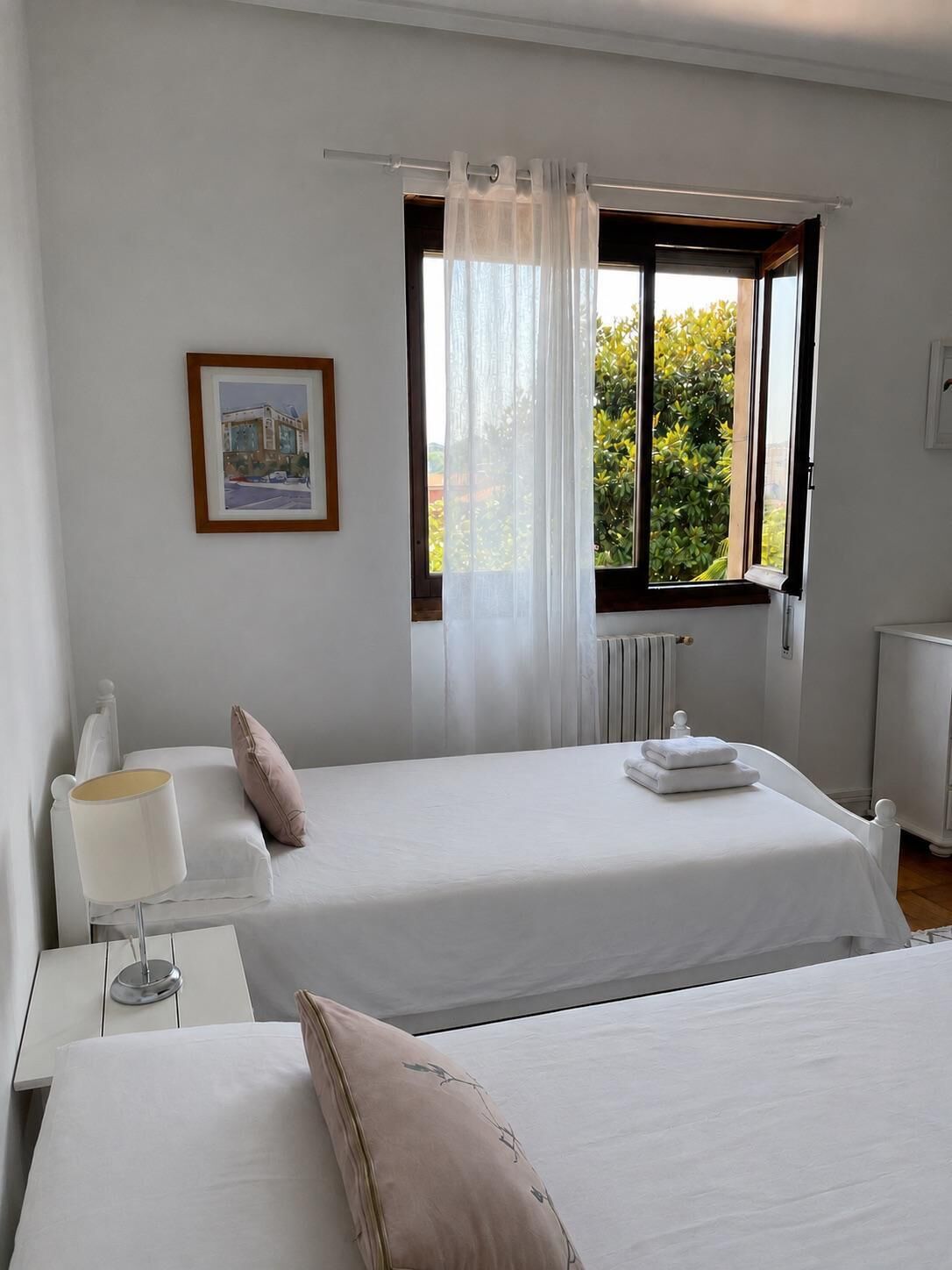 Habitación de hotel con dos camas, una ventana con cortinas, un radiador, una mesita de noche con lámpara y un cuadro enmarcado en la pared.