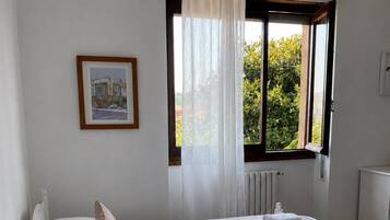 Habitación de hotel con dos camas, una ventana con cortinas, un radiador, una mesita de noche con lámpara y un cuadro enmarcado en la pared.