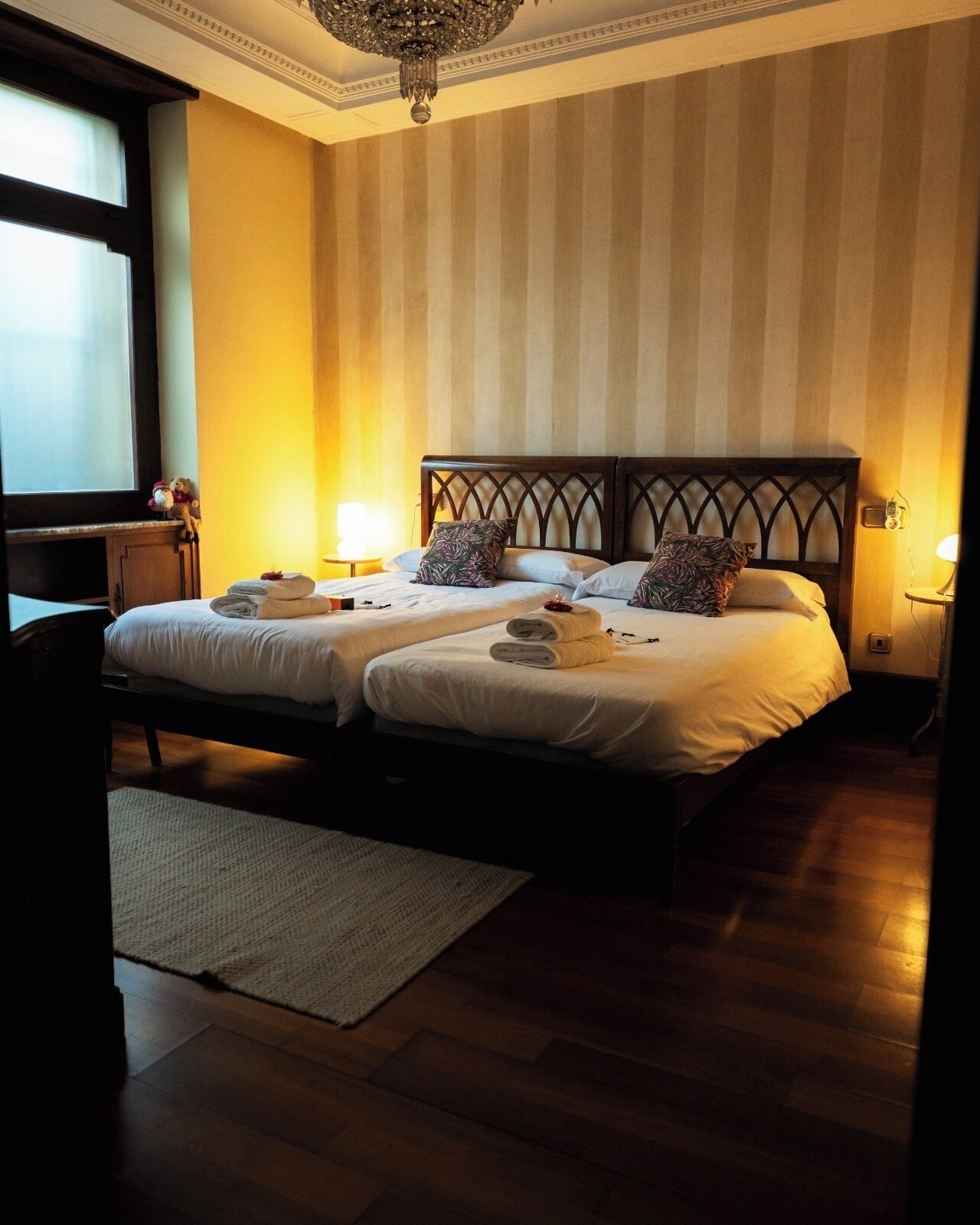 Ein Doppelbett mit Holz Kopfteil, ein Nachttisch mit Lampe und ein Fenster mit Vorhängen.