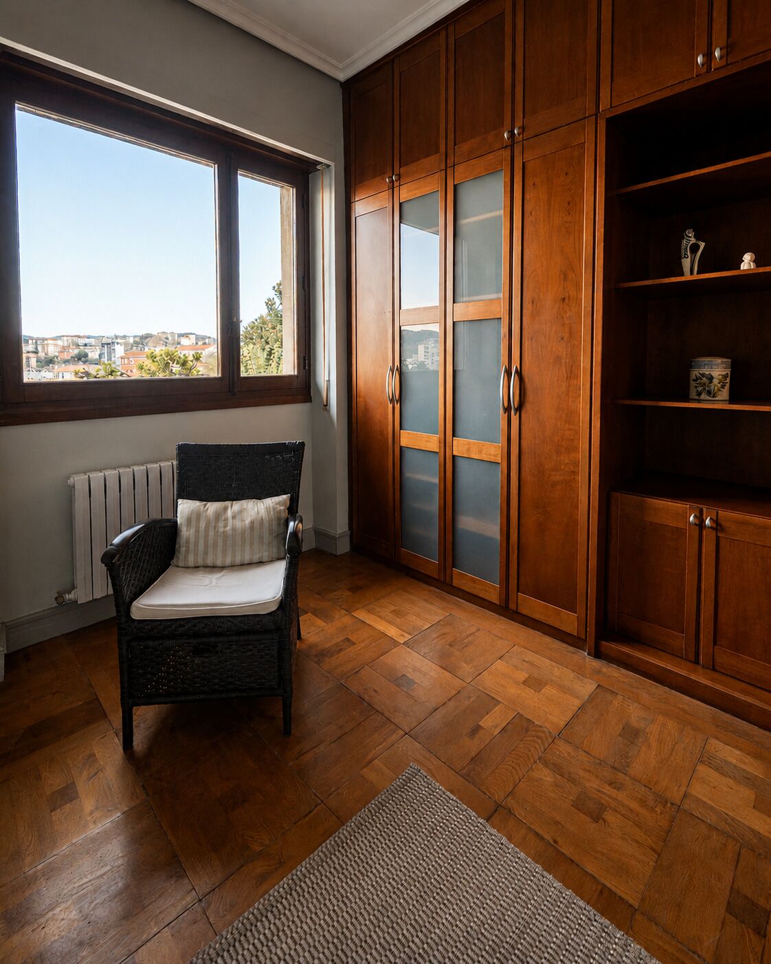 Una habitación con una silla de mimbre, armarios de madera, un radiador y vistas por una ventana.