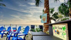 Rannan läheisyydessä sijaitseva oleskelualue, jossa on sinisiä tuoleja ja pöytä, palmu ja kyltti, jossa lukee: ”Tiki Time”.