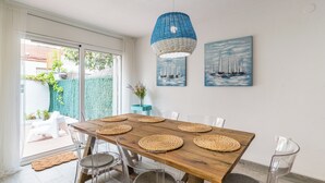 A dining area with a wooden table, clear chairs, woven placemats, a blue woven pendant light, and two paintings of sailboats.