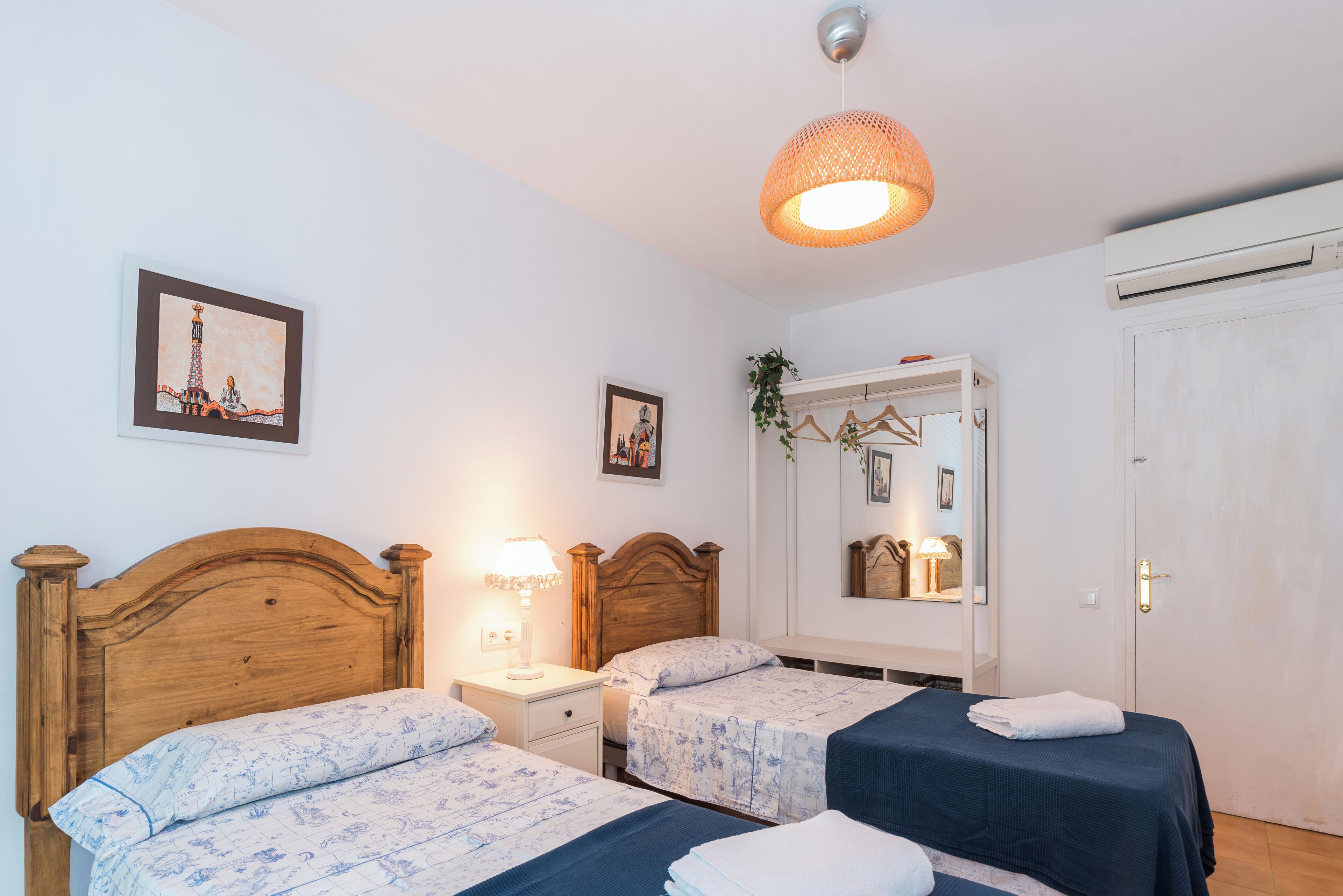 Una habitación con dos camas de madera, una mesita de noche con lámpara, una planta colgante, un espejo y una puerta.