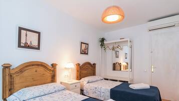 Una habitación con dos camas de madera, una mesita de noche con lámpara, una planta colgante, un espejo y una puerta.